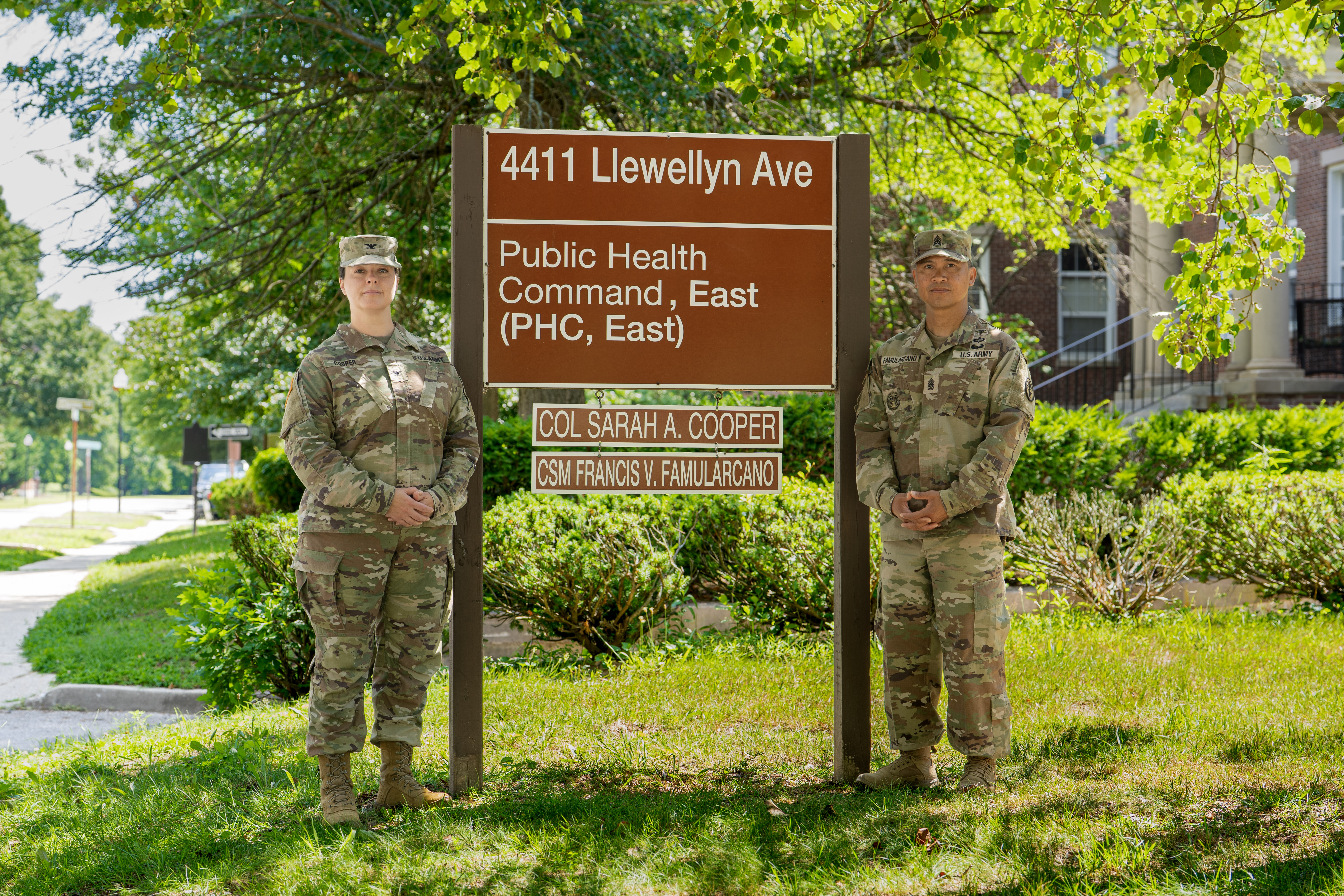 Two soldiers standing in front of building address sign, text is: "4411 Llewellyn Ave., Public Health Command, East, COL Sarah A. Cooper, CSM Francis V. Famularcano"